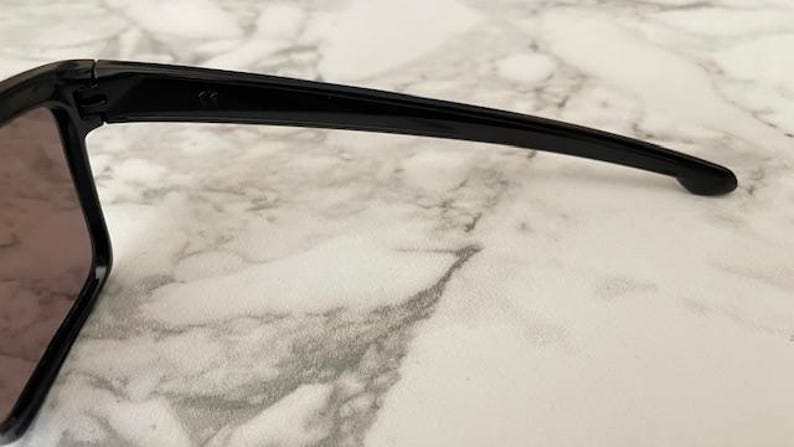 May include: Close-up of black sunglasses with a rectangular frame and dark lenses. The sunglasses have a sleek, modern design with a curved arm. The image is set against a marble-like background.