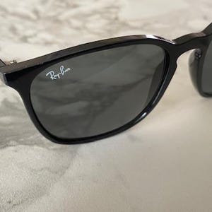 Puede incluir: Gafas de sol negras con lentes grises oscuras. El logotipo de Ray-Ban es visible en la lente derecha. Las gafas de sol tienen un acabado brillante y est&aacute;n sobre una superficie clara, similar al m&aacute;rmol.