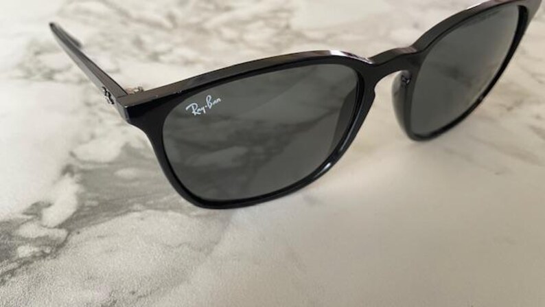 Puede incluir: Gafas de sol negras con lentes grises oscuras. El logotipo de Ray-Ban es visible en la lente derecha. Las gafas de sol tienen un acabado brillante y est&aacute;n sobre una superficie clara, similar al m&aacute;rmol.