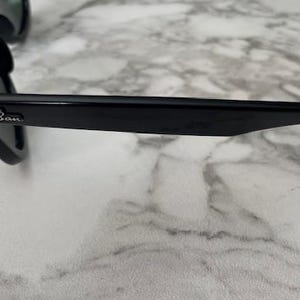 K&ouml;nnte beinhalten: Schwarze Sonnenbrille mit dunklen Gl&auml;sern. Der Rahmen ist gl&auml;nzend schwarz mit dem Ray-Ban-Logo am B&uuml;gel. Die Sonnenbrille liegt auf einer Marmoroberfl&auml;che.