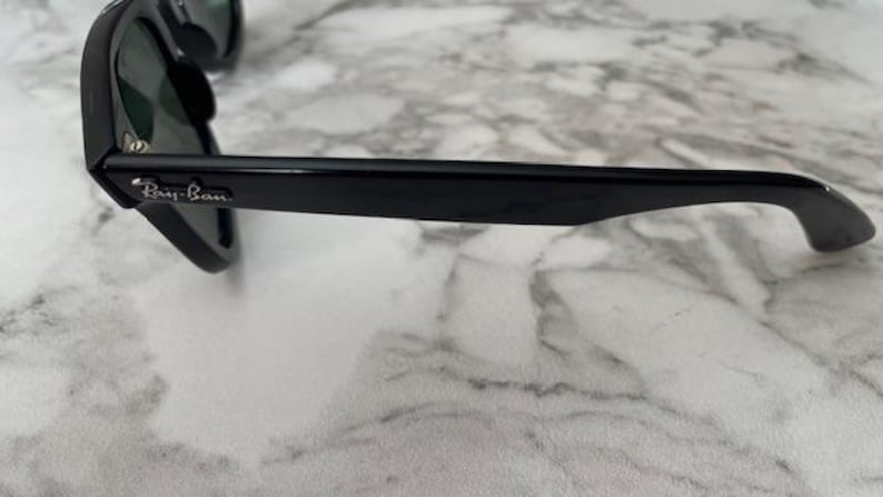 K&ouml;nnte beinhalten: Schwarze Sonnenbrille mit dunklen Gl&auml;sern. Der Rahmen ist gl&auml;nzend schwarz mit dem Ray-Ban-Logo am B&uuml;gel. Die Sonnenbrille liegt auf einer Marmoroberfl&auml;che.