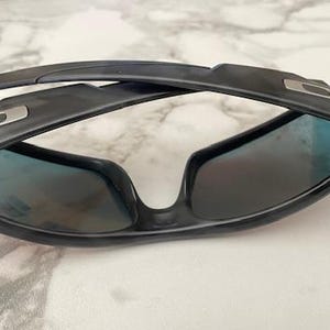 Puede incluir: Gafas de sol negras con lentes tintadas en azul. Las gafas tienen un dise&ntilde;o elegante y moderno con montura y patillas negras. Las lentes son ligeramente reflectantes y la montura presenta un detalle plateado.