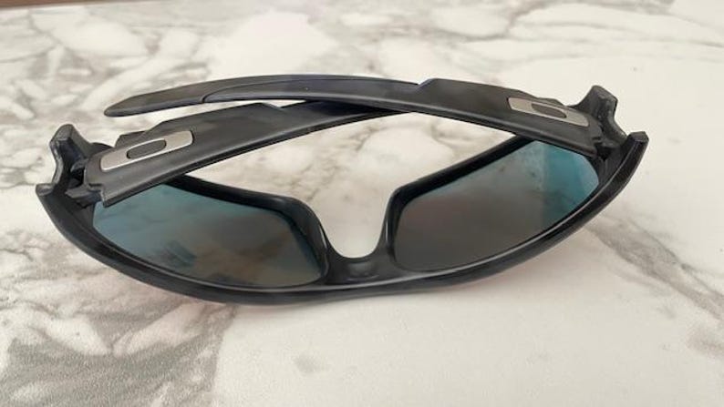 Puede incluir: Gafas de sol negras con lentes tintadas en azul. Las gafas tienen un dise&ntilde;o elegante y moderno con montura y patillas negras. Las lentes son ligeramente reflectantes y la montura presenta un detalle plateado.