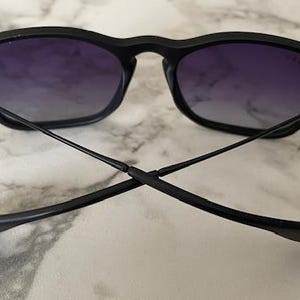 May include: Black sunglasses with a square frame and gradient purple lenses. The temples are thin and extend outward, crossing each other. The sunglasses are set against a marble background.