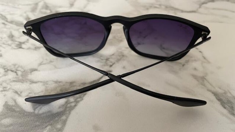 May include: Black sunglasses with a square frame and gradient purple lenses. The temples are thin and extend outward, crossing each other. The sunglasses are set against a marble background.