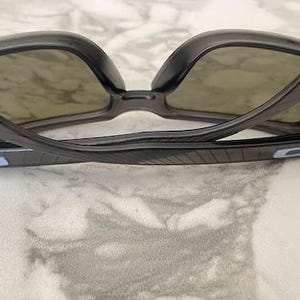 May include: Dark gray sunglasses with a curved frame and tinted lenses. The temples have a blue rectangular accent. The sunglasses are positioned on a light-colored surface, showcasing their design and shape.