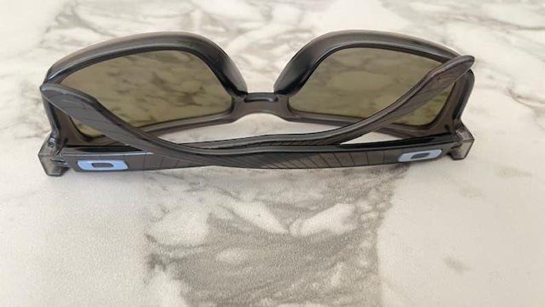 May include: Dark gray sunglasses with a curved frame and tinted lenses. The temples have a blue rectangular accent. The sunglasses are positioned on a light-colored surface, showcasing their design and shape.