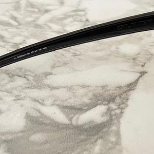 May include: Close-up of black sunglasses with dark lenses. The arm of the sunglasses has the text "FL 00934-02 57 18 140". The sunglasses are set against a marble-like background.