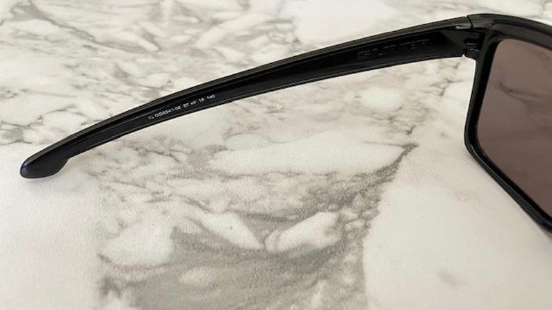 May include: Close-up of black sunglasses with dark lenses. The arm of the sunglasses has the text "FL 00934-02 57 18 140". The sunglasses are set against a marble-like background.