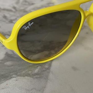 May include: Bright yellow sunglasses with dark lenses. The Ray-Ban logo is visible on the lens. The sunglasses have a thick, rounded frame and arms. The glasses are set against a light-colored background.