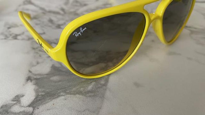 May include: Bright yellow sunglasses with dark lenses. The Ray-Ban logo is visible on the lens. The sunglasses have a thick, rounded frame and arms. The glasses are set against a light-colored background.