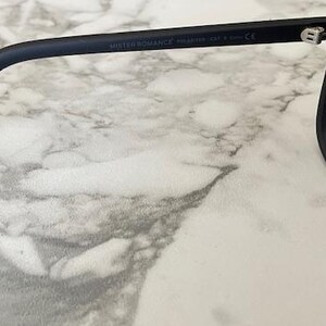 Puede incluir: Gafas de sol en negro mate con lentes oscuras y una patilla curva. El interior de la patilla muestra el texto MISTER ROMANCE, seguido de la palabra POLARIZED y la marca CE, lo que indica el cumplimiento de las normas de seguridad.