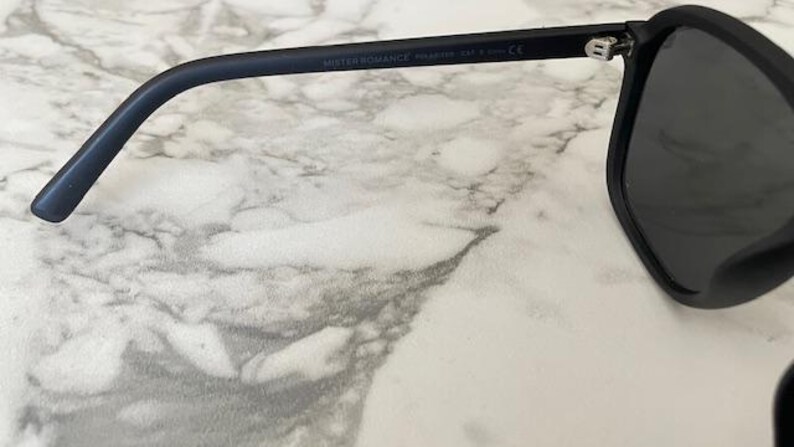 Puede incluir: Gafas de sol en negro mate con lentes oscuras y una patilla curva. El interior de la patilla muestra el texto MISTER ROMANCE, seguido de la palabra POLARIZED y la marca CE, lo que indica el cumplimiento de las normas de seguridad.