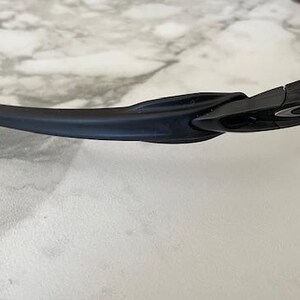 Puede incluir: Gafas de sol negras con lentes amarillas. Las gafas de sol tienen un dise&ntilde;o elegante y moderno con una montura negra y una patilla curvada. El logotipo de Oakley es visible en la montura.