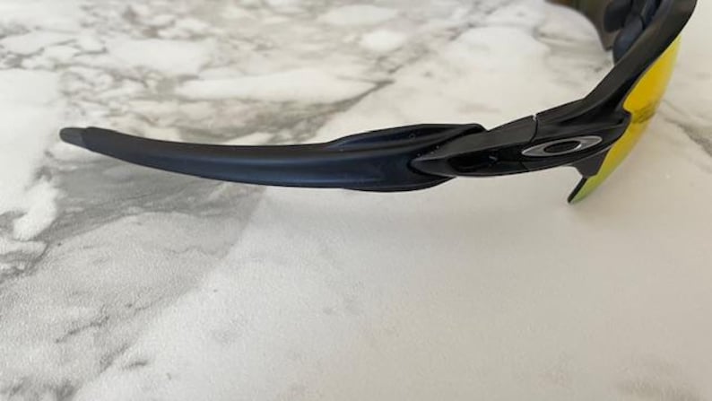 Puede incluir: Gafas de sol negras con lentes amarillas. Las gafas de sol tienen un dise&ntilde;o elegante y moderno con una montura negra y una patilla curvada. El logotipo de Oakley es visible en la montura.