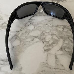 May include: Black sunglasses with a rectangular frame and dark lenses. The sunglasses have a curved design and are set against a marble background. The arms of the sunglasses are long and thin.