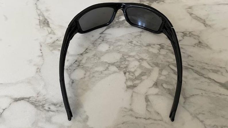 May include: Black sunglasses with a rectangular frame and dark lenses. The sunglasses have a curved design and are set against a marble background. The arms of the sunglasses are long and thin.