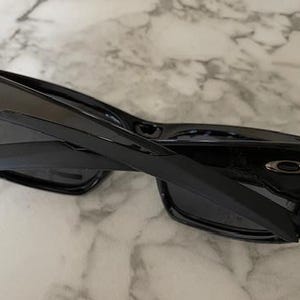 May include: Black sunglasses with a glossy finish. The sunglasses have a rectangular frame and dark lenses. The arms are thick and feature a silver logo. The sunglasses are folded.