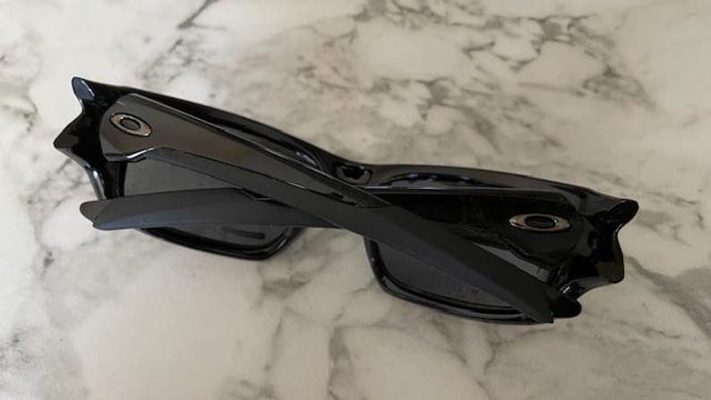 May include: Black sunglasses with a glossy finish. The sunglasses have a rectangular frame and dark lenses. The arms are thick and feature a silver logo. The sunglasses are folded.