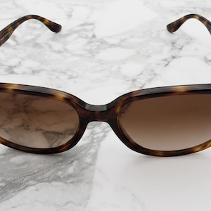 May include: Tortoiseshell sunglasses with brown gradient lenses. The Ray-Ban logo is visible on the left lens. The frame has a classic, rounded shape and is set against a white marble background.