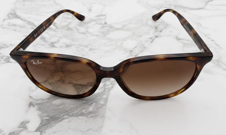 May include: Tortoiseshell sunglasses with brown gradient lenses. The Ray-Ban logo is visible on the left lens. The frame has a classic, rounded shape and is set against a white marble background.