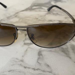 May include: Sunglasses with a dark metal frame and gradient brown lenses. The sunglasses have a curved design and a small logo on the side of the frame. The lenses transition from a darker shade at the top to a lighter shade at the bottom.