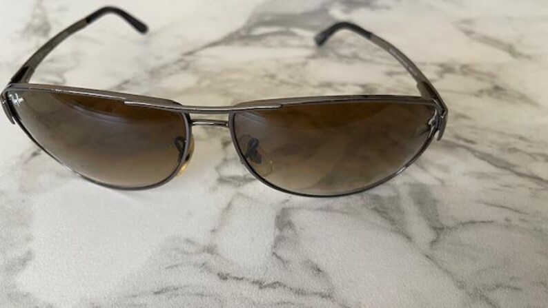 May include: Sunglasses with a dark metal frame and gradient brown lenses. The sunglasses have a curved design and a small logo on the side of the frame. The lenses transition from a darker shade at the top to a lighter shade at the bottom.