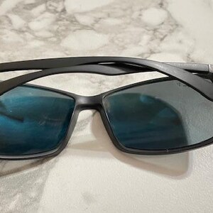 May include: Black sunglasses with rectangular frames and blue-tinted lenses. The sunglasses have a sleek, modern design, with a matte black finish on the frames and arms. The lenses reflect light, creating a blue hue.