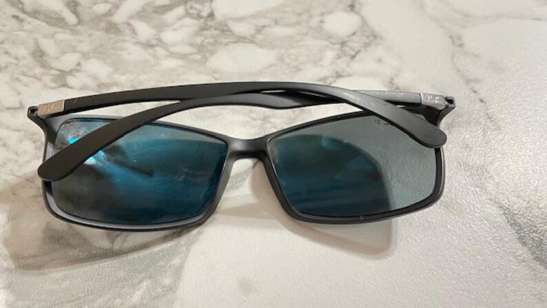 May include: Black sunglasses with rectangular frames and blue-tinted lenses. The sunglasses have a sleek, modern design, with a matte black finish on the frames and arms. The lenses reflect light, creating a blue hue.