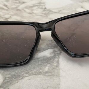 May include: Black sunglasses with a glossy finish. The sunglasses have a rectangular frame and dark lenses. The image is a close-up shot of the sunglasses, showcasing their design and shape. The sunglasses are resting on a light-colored surface.