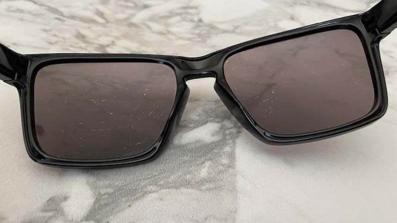 May include: Black sunglasses with a glossy finish. The sunglasses have a rectangular frame and dark lenses. The image is a close-up shot of the sunglasses, showcasing their design and shape. The sunglasses are resting on a light-colored surface.