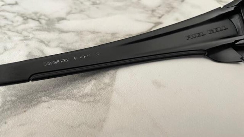 May include: Close-up of a black sunglasses arm. The text "FUEL CELL" is visible on the arm. The model number "OO9096-K66" and other markings are also present. The sunglasses arm is made of a smooth, matte material.
