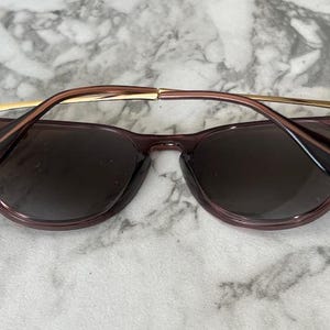 May include: Sunglasses with a dark purple frame and lenses. The temples are a light brown color with gold accents. The sunglasses are resting on a white and gray marble surface.