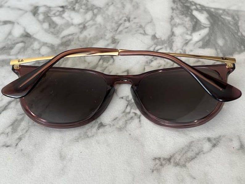 May include: Sunglasses with a dark purple frame and lenses. The temples are a light brown color with gold accents. The sunglasses are resting on a white and gray marble surface.
