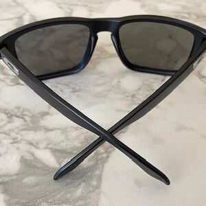 May include: Black sunglasses with dark gray lenses. The sunglasses have a rectangular frame and straight arms that cross at the ends. The glasses are set against a marble background.