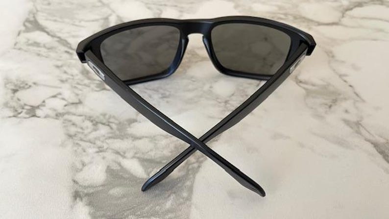 May include: Black sunglasses with dark gray lenses. The sunglasses have a rectangular frame and straight arms that cross at the ends. The glasses are set against a marble background.