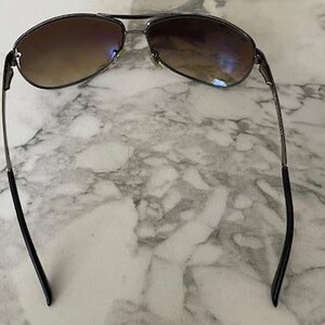May include: Sunglasses with a dark metal frame and gradient brown lenses. The lenses transition from a darker shade at the top to a lighter shade at the bottom. The temples are thin and black, with a slight curve.