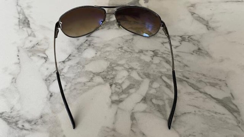 May include: Sunglasses with a dark metal frame and gradient brown lenses. The lenses transition from a darker shade at the top to a lighter shade at the bottom. The temples are thin and black, with a slight curve.