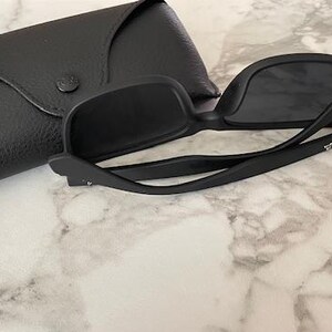 Pu&ograve; includere: Occhiali da sole neri con custodia nera abbinata. Gli occhiali hanno una montatura quadrata e lenti scure. La custodia &egrave; realizzata in materiale nero strutturato e presenta un logo dorato. Gli occhiali sono aperti, mostrando la montatura e le lenti.