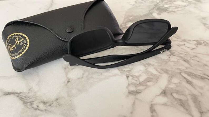 Pu&ograve; includere: Occhiali da sole neri con custodia nera abbinata. Gli occhiali hanno una montatura quadrata e lenti scure. La custodia &egrave; realizzata in materiale nero strutturato e presenta un logo dorato. Gli occhiali sono aperti, mostrando la montatura e le lenti.