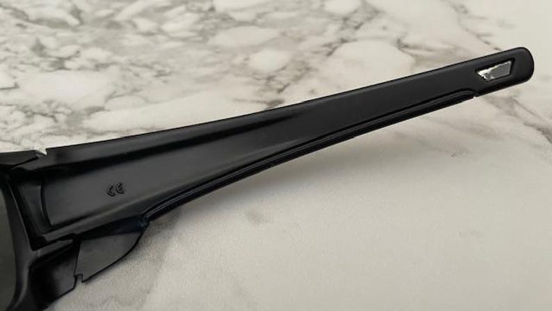May include: Close-up of a black sunglasses arm. The arm is sleek and glossy, with a small, reflective silver accent near the temple. The frame has a subtle curve and a CE marking.