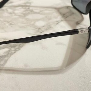 May include: Black sunglasses with a sleek, modern design. The sunglasses have dark lenses and a black frame with a silver accent near the temple. The Ray-Ban logo is visible on the frame.