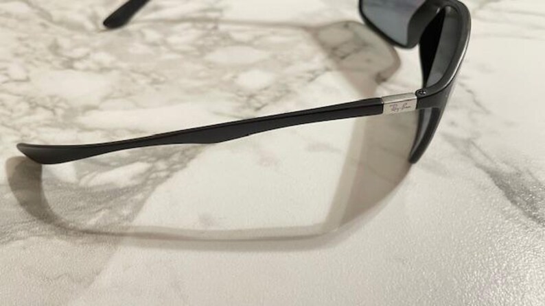 May include: Black sunglasses with a sleek, modern design. The sunglasses have dark lenses and a black frame with a silver accent near the temple. The Ray-Ban logo is visible on the frame.