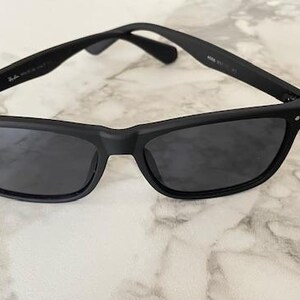 Pu&ograve; includere: Occhiali da sole neri con finitura opaca. Gli occhiali hanno una montatura rettangolare con lenti scure e il logo Ray-Ban sulla lente sinistra. Le aste sono sottili e la montatura ha piccoli accenti argentati.