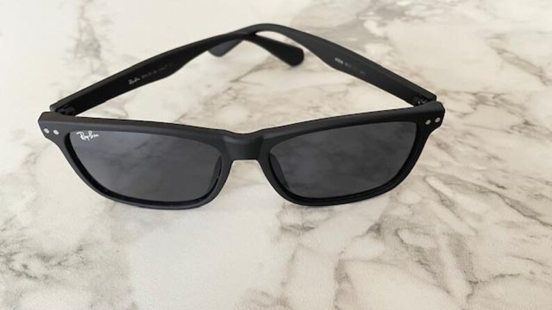 Pu&ograve; includere: Occhiali da sole neri con finitura opaca. Gli occhiali hanno una montatura rettangolare con lenti scure e il logo Ray-Ban sulla lente sinistra. Le aste sono sottili e la montatura ha piccoli accenti argentati.
