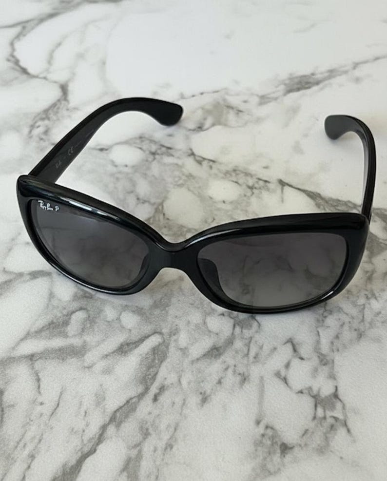 K&ouml;nnte beinhalten: Schwarze Sonnenbrille mit geschwungenem Rahmen und dunklen Gl&auml;sern. Die Sonnenbrille hat eine subtile Cat-Eye-Form und das Ray-Ban-Logo ist auf dem linken Glas sichtbar. Auf einem Marmorhintergrund.