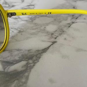 May include: Bright yellow sunglasses with dark lenses. The frame is a vibrant yellow, and the arm of the glasses has the text "MADE IN ITALY" and the Ray-Ban logo. The glasses are set against a marble background.