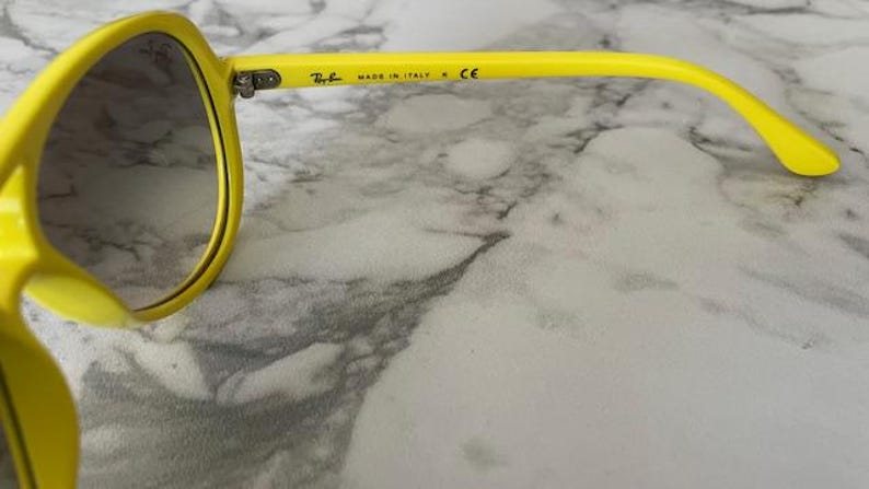 May include: Bright yellow sunglasses with dark lenses. The frame is a vibrant yellow, and the arm of the glasses has the text "MADE IN ITALY" and the Ray-Ban logo. The glasses are set against a marble background.