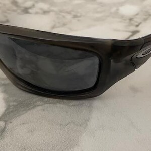 May include: Close-up of black sunglasses with dark lenses. The sunglasses have a thick, rectangular frame and a small silver emblem on the side. The lenses are reflective, and the sunglasses are resting on a marble surface.