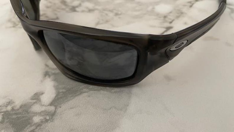May include: Close-up of black sunglasses with dark lenses. The sunglasses have a thick, rectangular frame and a small silver emblem on the side. The lenses are reflective, and the sunglasses are resting on a marble surface.